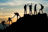 Silhouette of a group of people climbing a mountain, helping each other, with a sunset in the background symbolising teamwork and achievement.